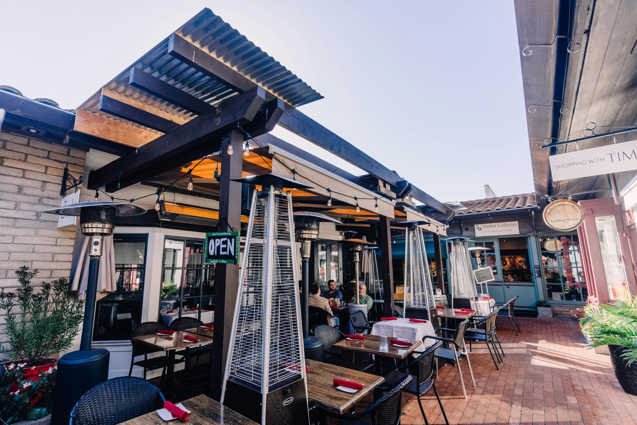 Sunny outdoor restaurant setting with tables, chairs, and heaters under a modern pergola.