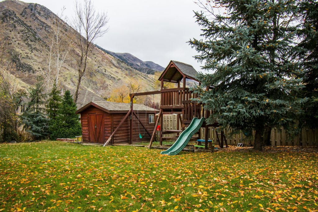 A serene autumn scene featuring a wooden playground and shed in a backyard with fallen leaves.