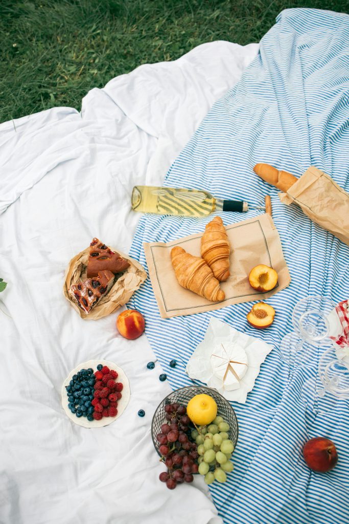 A beautifully arranged summer picnic with croissants, fresh fruits, and white wine on a blanket.
