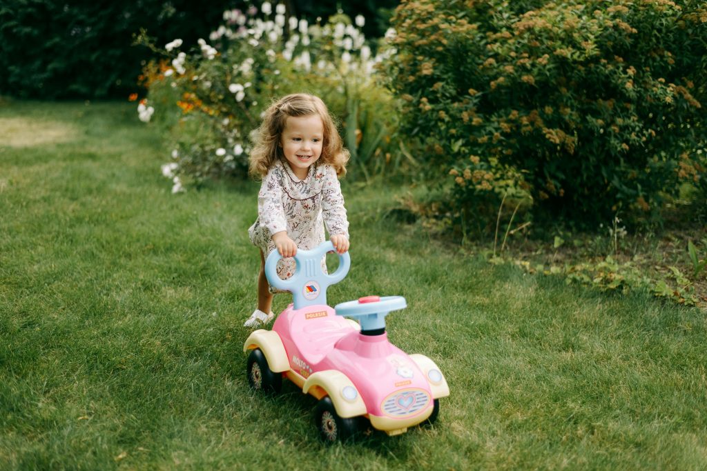 A joyful toddler girl in a floral dress plays with a pink toy car on a sunny day in the garden.