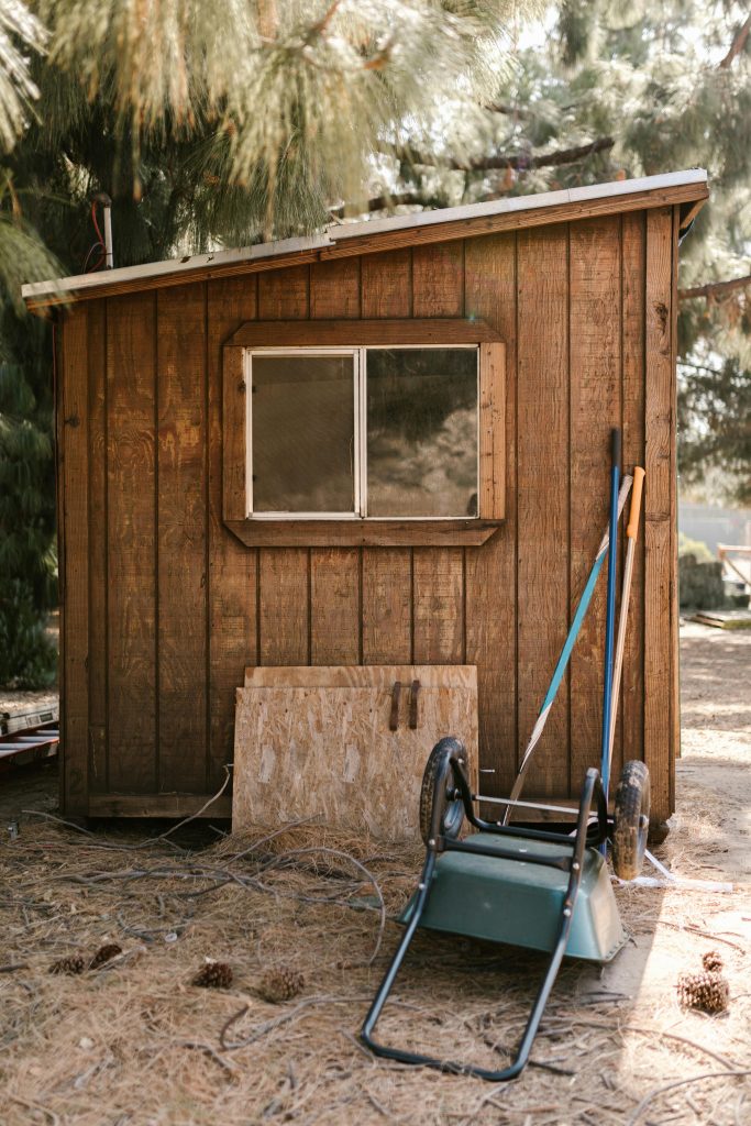 A rustic wooden shed surrounded by farm tools and pine trees, embodying rural simplicity.