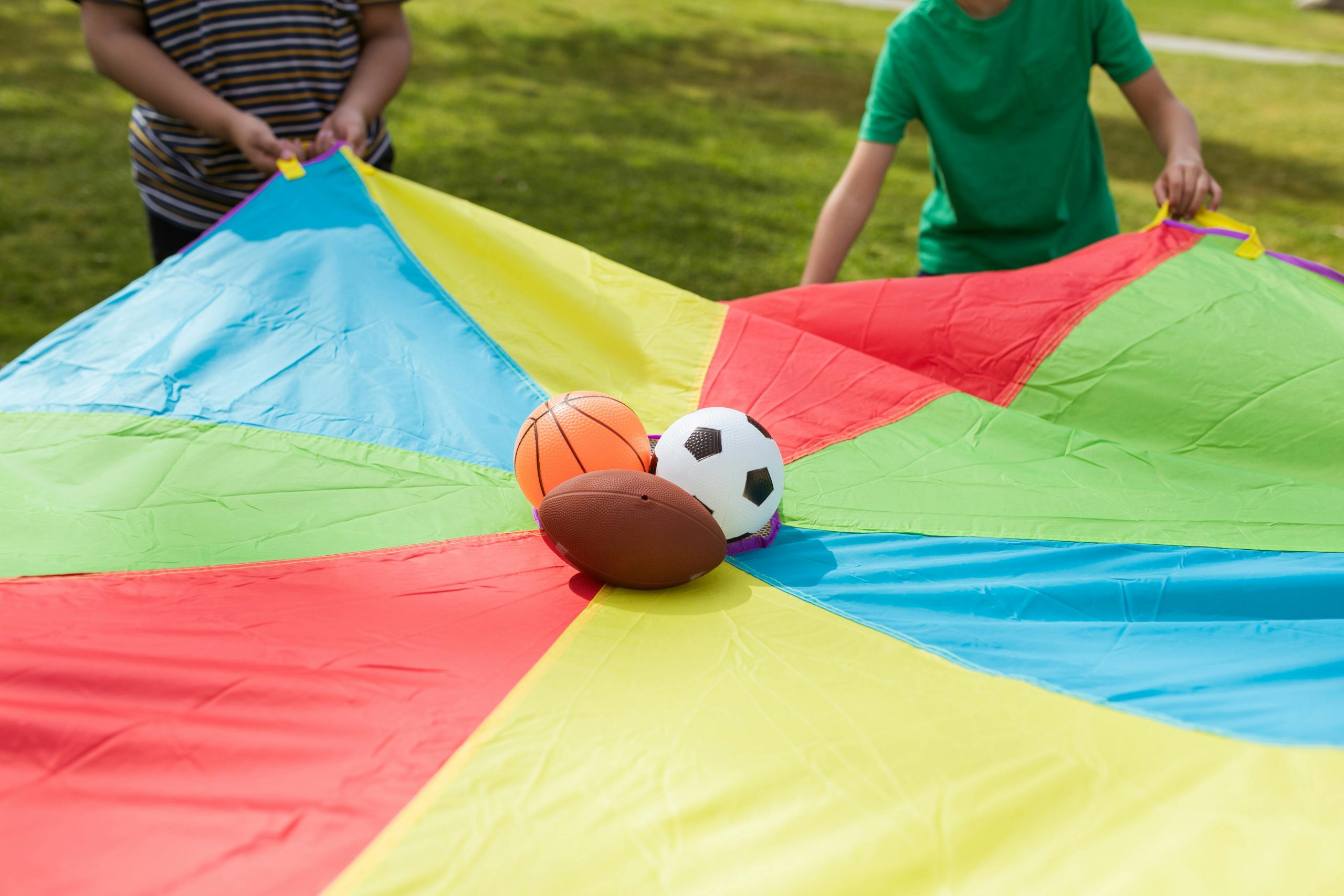 Children playing outside with a colorful parachute and sports balls on grass.